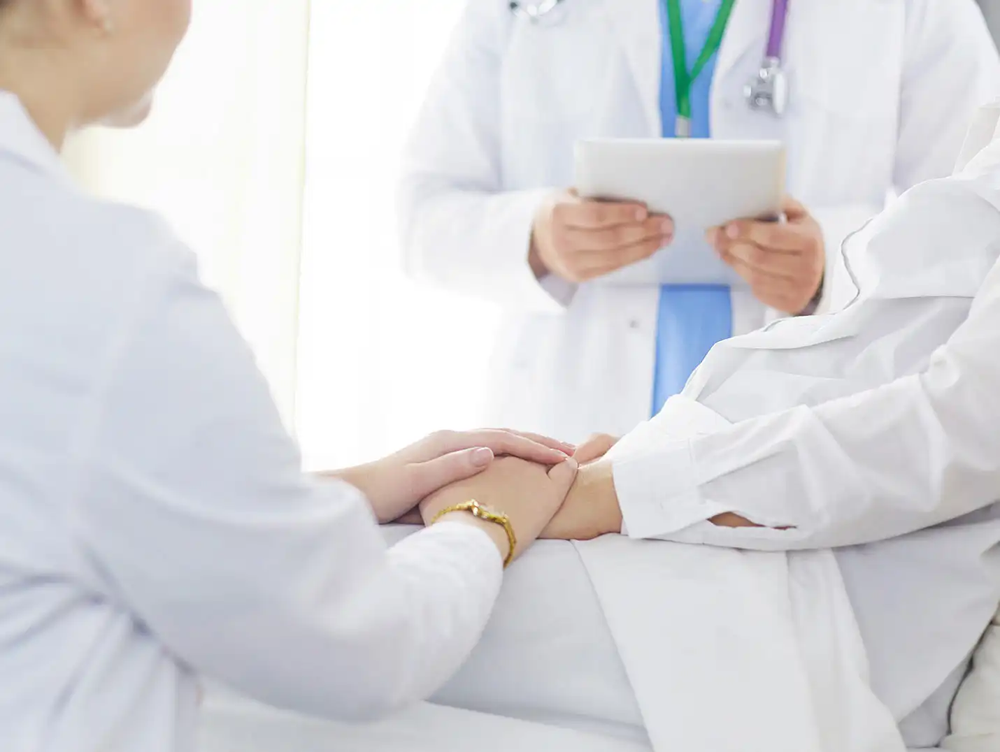 Doctora sentada y colocando sus manos sobre las de un paciente que está recostado. Otro doctor está 
sosteniendo un iPad