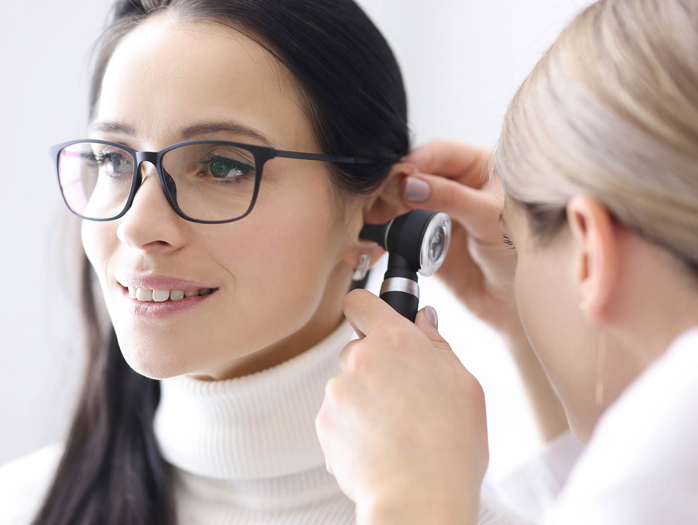 Doctora revisando con un otoscopio el oído de una paciente que está vestida con un suéter blanco y usando lentes negros