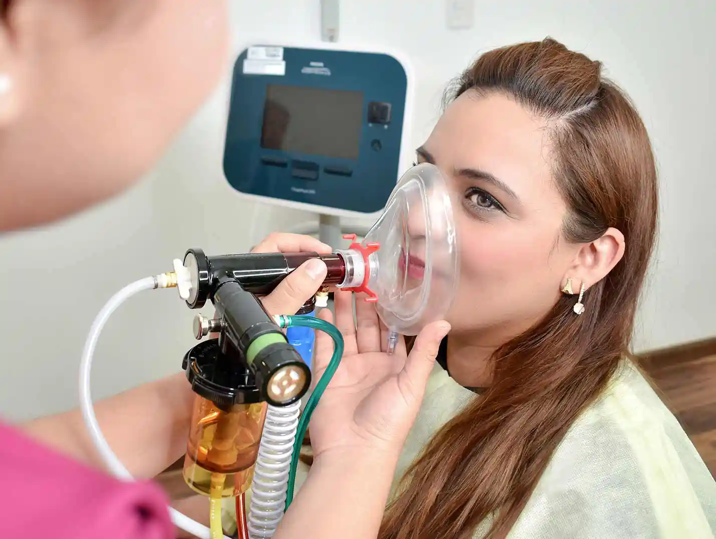 Doctora colocando una mascarilla de oxígeno a una paciente que la mira detenidamente. A su lado hay un monitor blanco