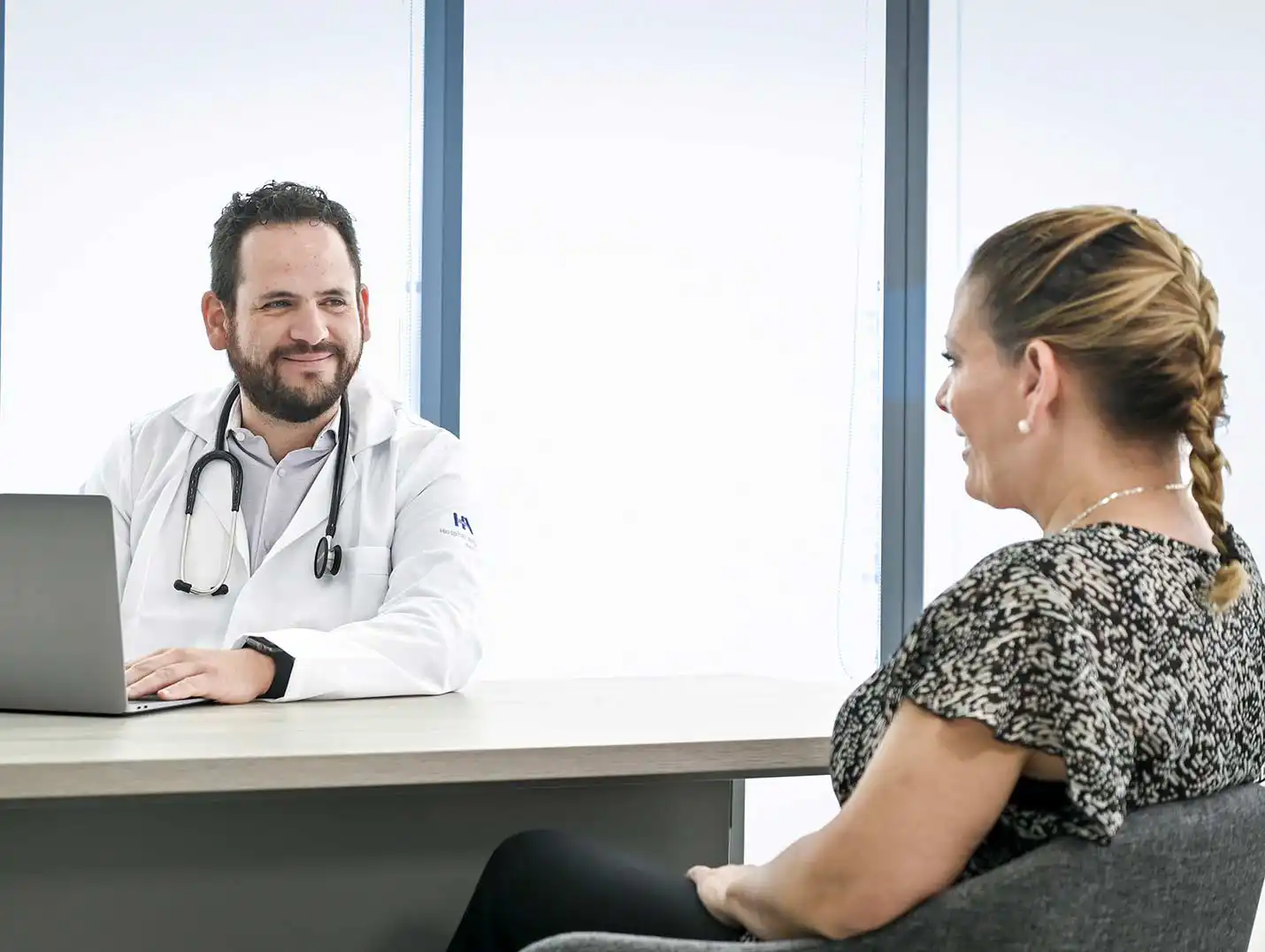 Doctor con sus manos sobre el teclado de una MacBook, mientras escucha y mira a su paciente que está sentada frente a él