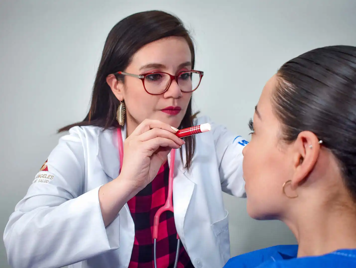 Doctora de Hospital Angeles usando una bata blanca sobre una blusa de cuadros, evaluando a una paciente con una lámpara