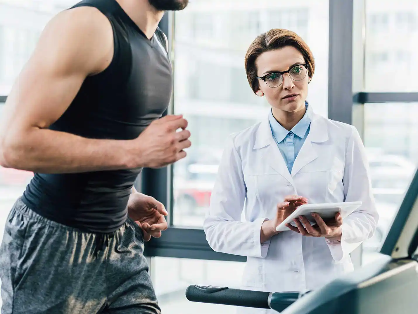 Paciente vestido con una playera deportiva negra y pants gris, corriendo en una caminadora mientras una doctora lo monitorea