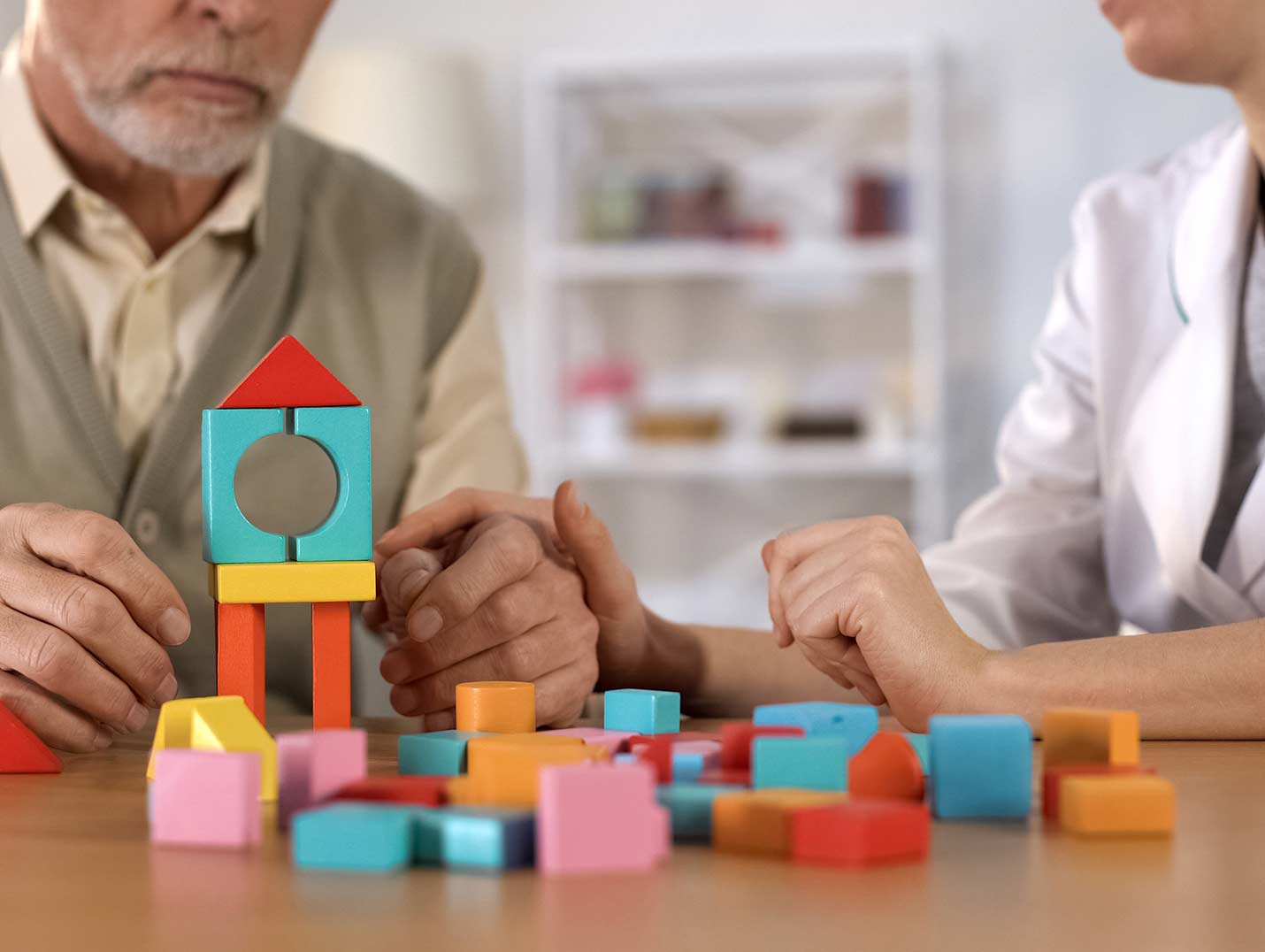 Paciente de la tercera edad acomodando bloques de juguete azule, rojos, naranjas, amarillos y rosas. A su lado está una doctora