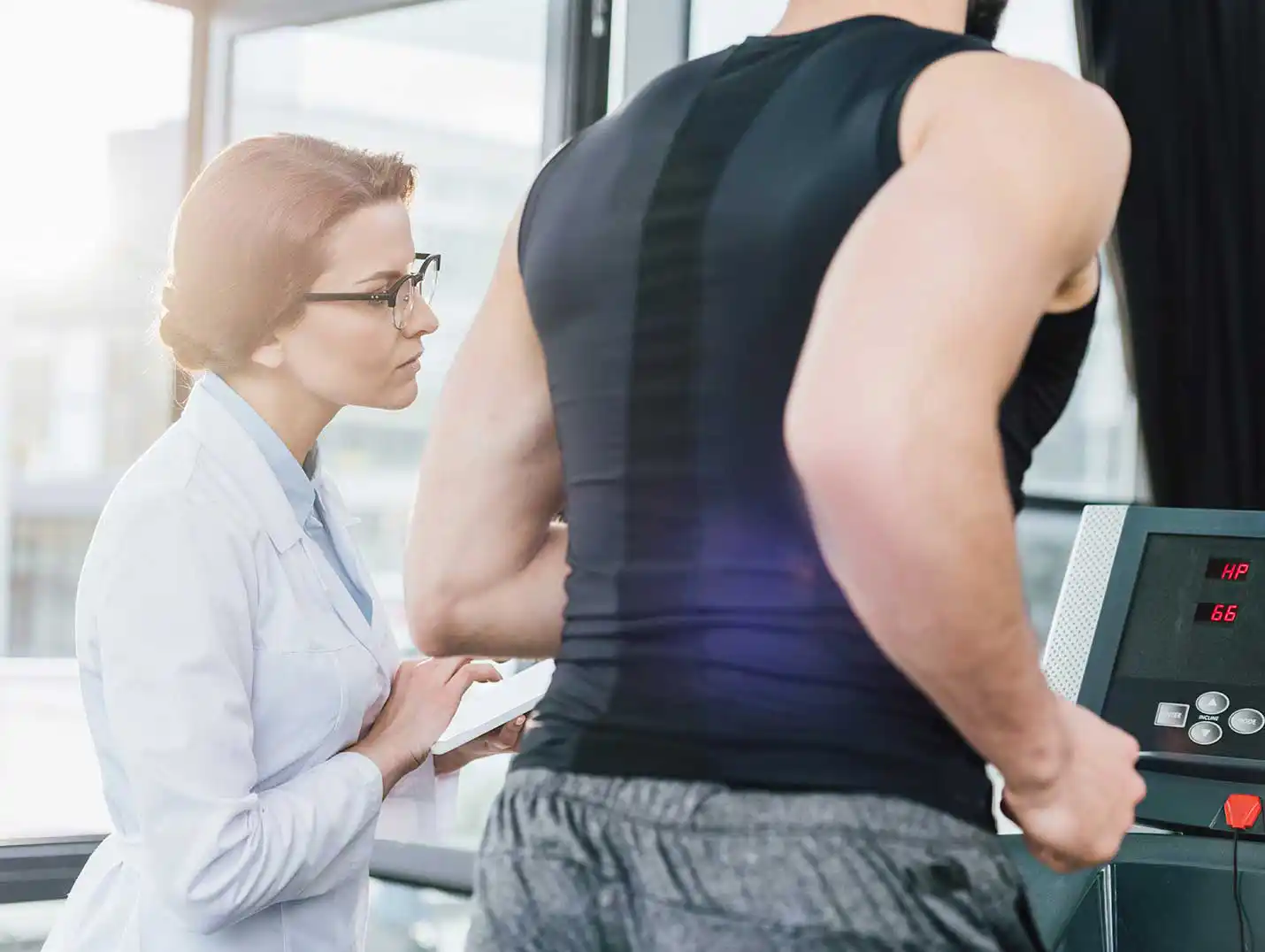 Paciente vestido con una playera deportiva negra y pants gris, corriendo en una caminadora mientras una doctora lo monitorea