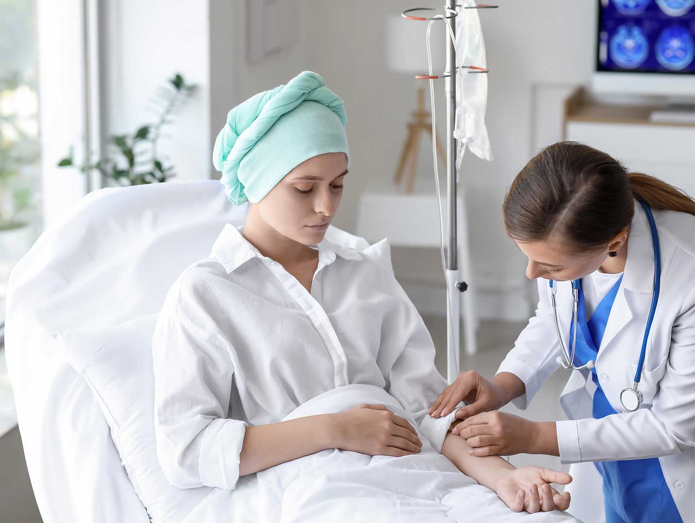 Paciente oncológico con playera blanca y mascada verde en la cabeza recostado sobre la cama siendo revisado por un especialista.