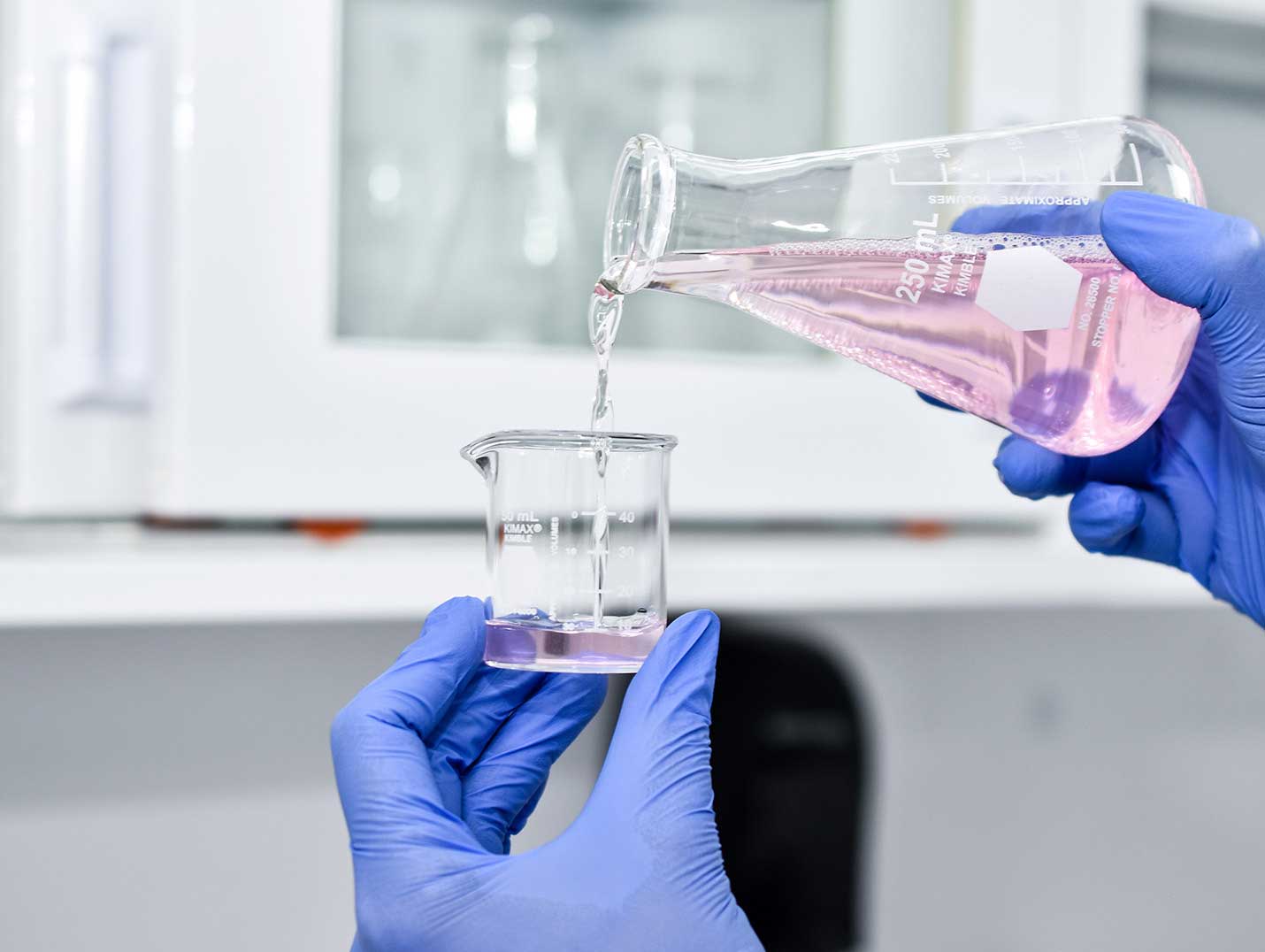Manos de un médico con guantes azules, sosteniendo un matraz y vaciando una solución rosa en un vaso precipitado de vidrio