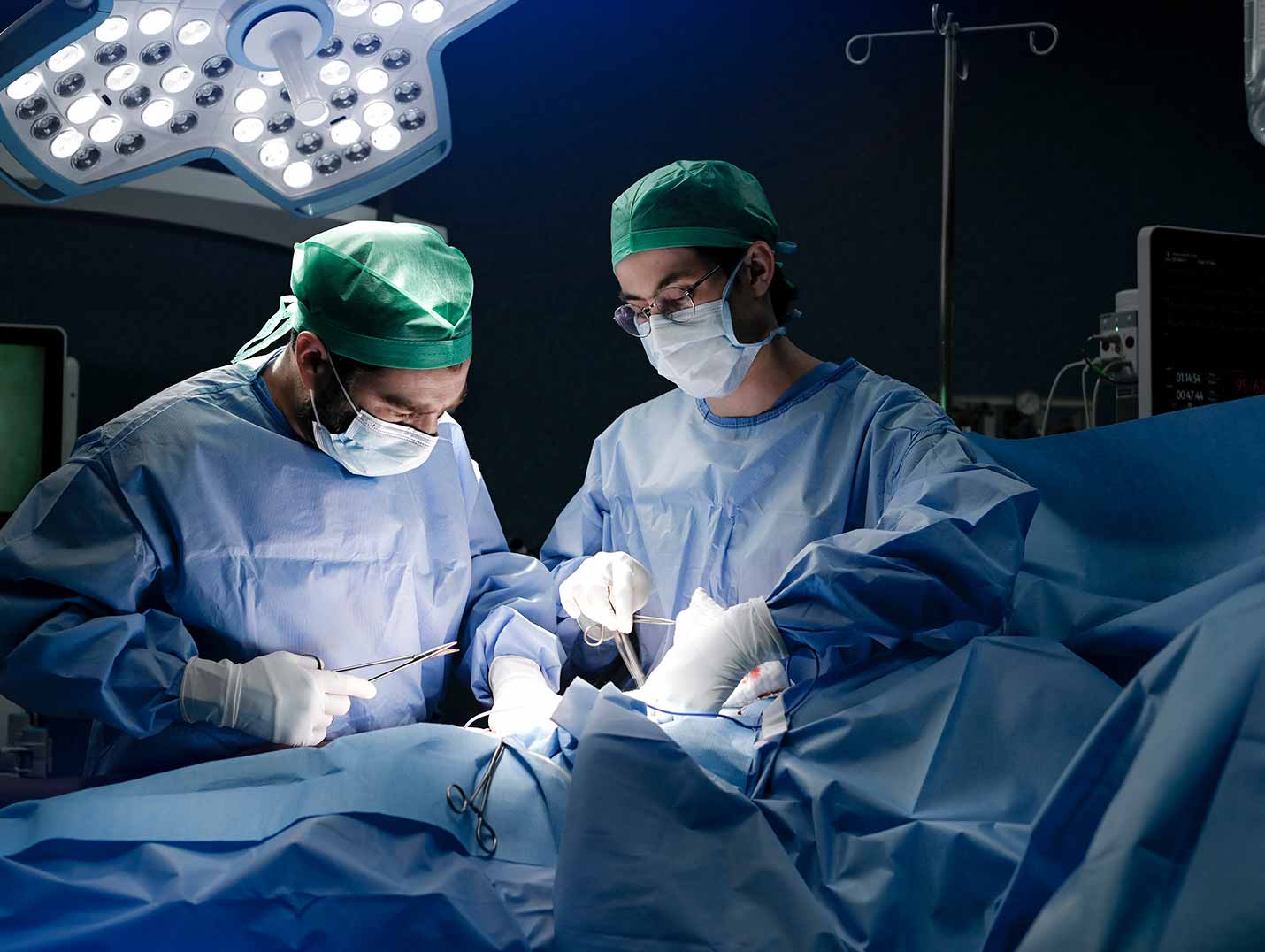 Dos médicos con pijamas azules para cirugía, gorros y cubrebocas realizando un procedimiento quirúrgico
