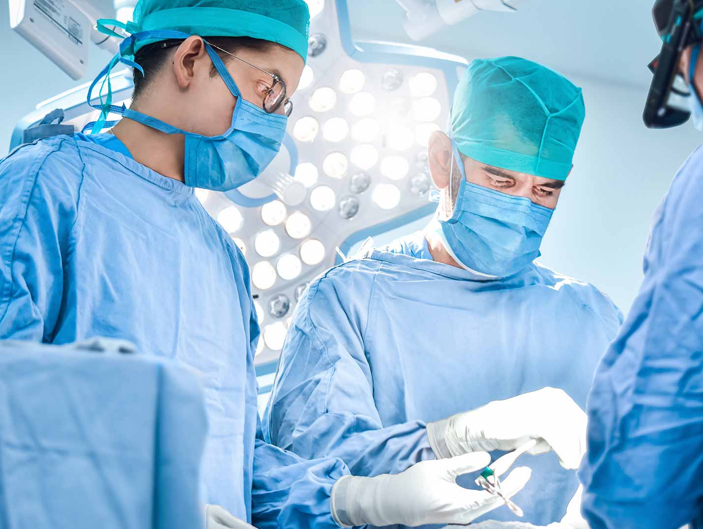 Tres médicos con pijamas azules para cirugía, gorros y cubrebocas realizando un procedimiento quirúrgico