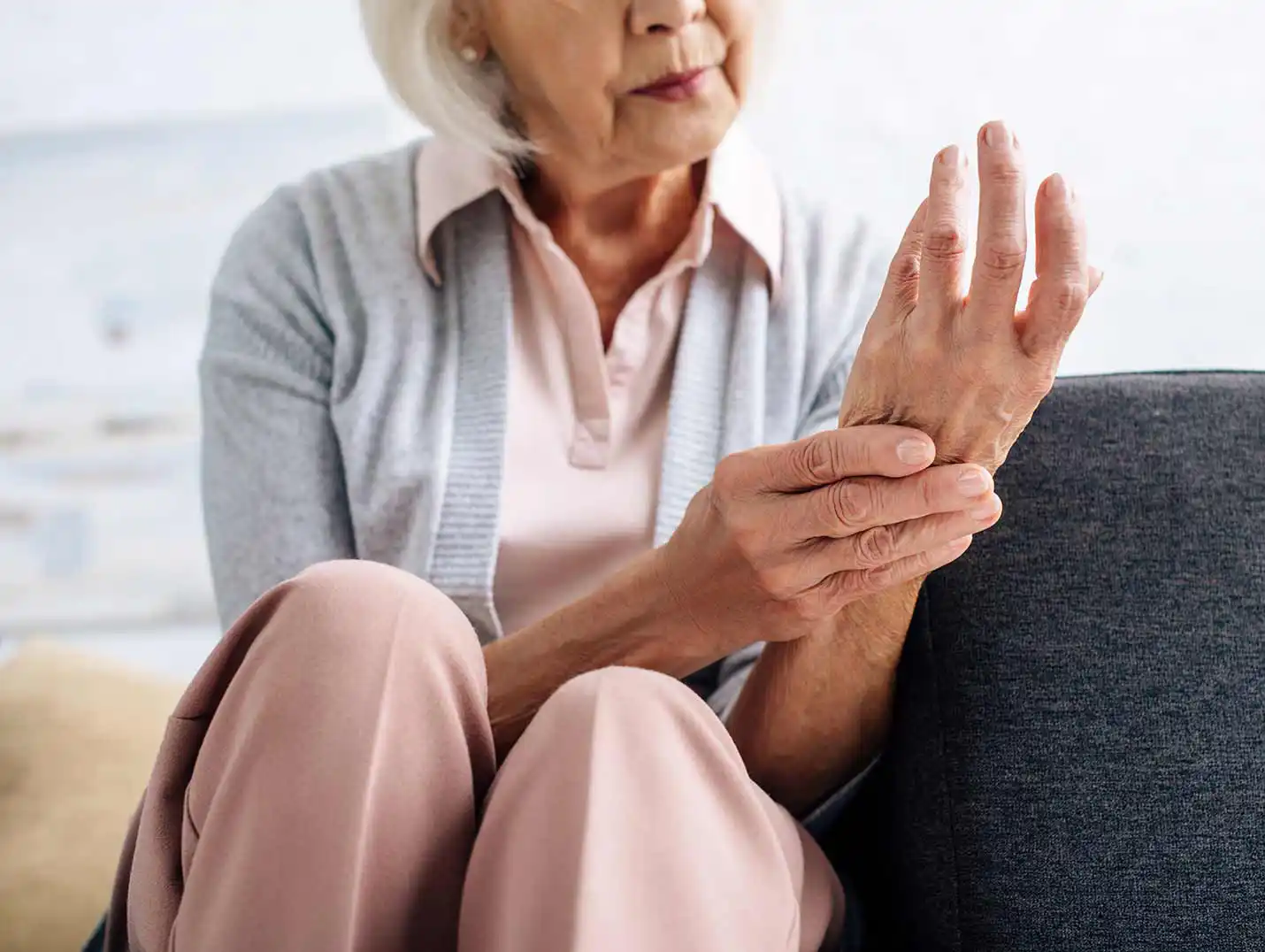 Mujer de la tercera edad sentada, vestida con una blusa y pantalón rosa y un suéter gris, agarrando su mano izquierda