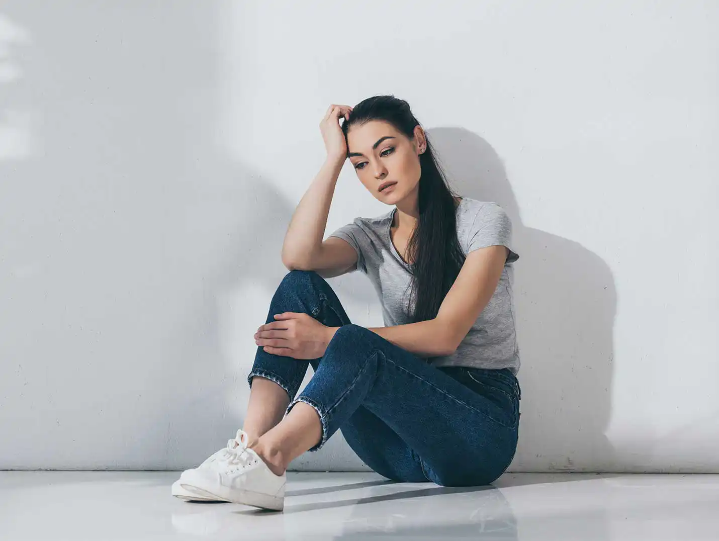 Mujer joven pensativa y sentada en el suelo de un cuarto amplio, vestida con una playera gris, pantalón de mezclilla y tenis