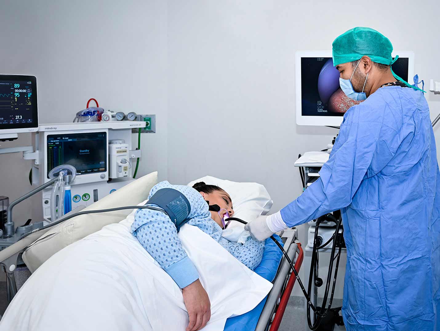 Doctor con pijama azul para cirugía, gorro verde y guantes blancos, realizando una endoscopia digestiva a una paciente