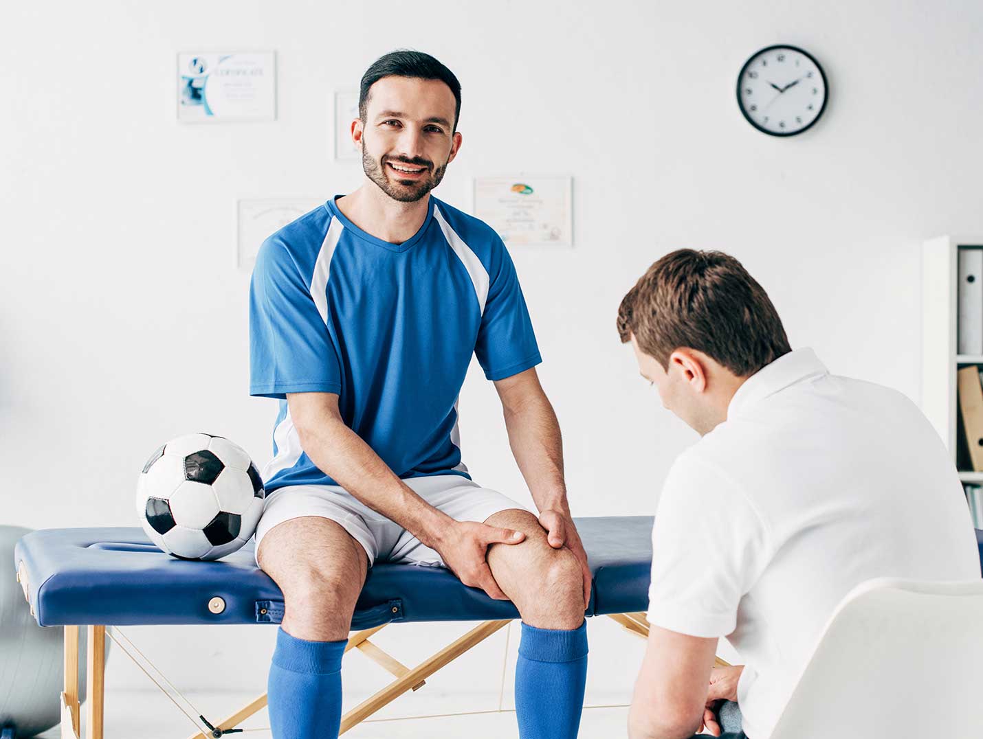 Futbolista con uniforme azul y blanco, sentado en una camilla terapéutica azul, mientras un doctor ve su rodilla