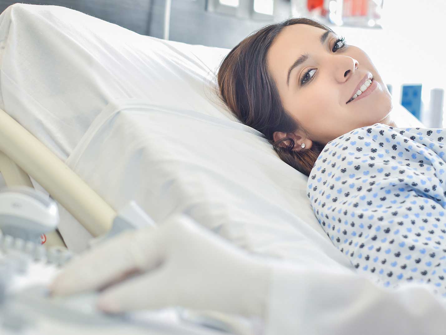 Mano del médico con un guante blanco sobre un monitor. A su lado está una paciente acostada en una cama hospitalaria