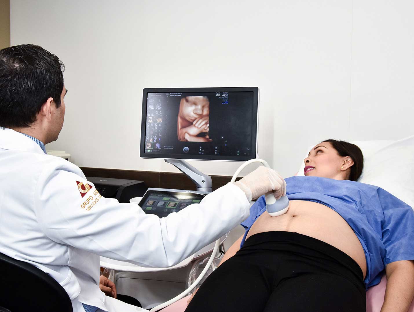 Médico realizando un ultrasonido a una paciente embarazada que está recostada. Ambos ven al monitor que muestra al bebé