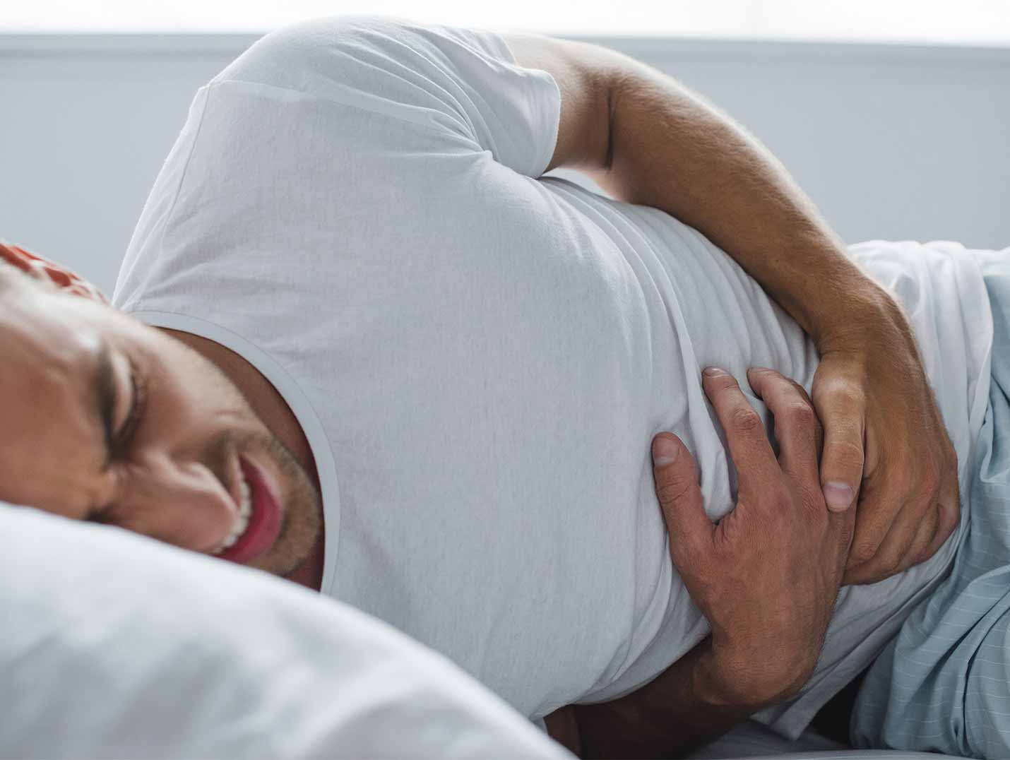 Hombre joven vistiendo una playera blanca, acostado en una cama, tocando su estómago y quejándose de dolor