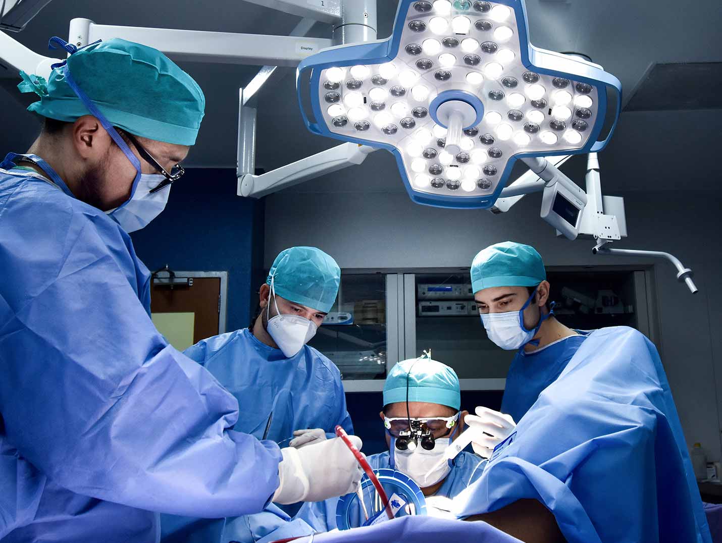 Tres médicos vestidos con pijamas azules para cirugía, gorros y cubrebocas, realizando una cirugía en un quirófano