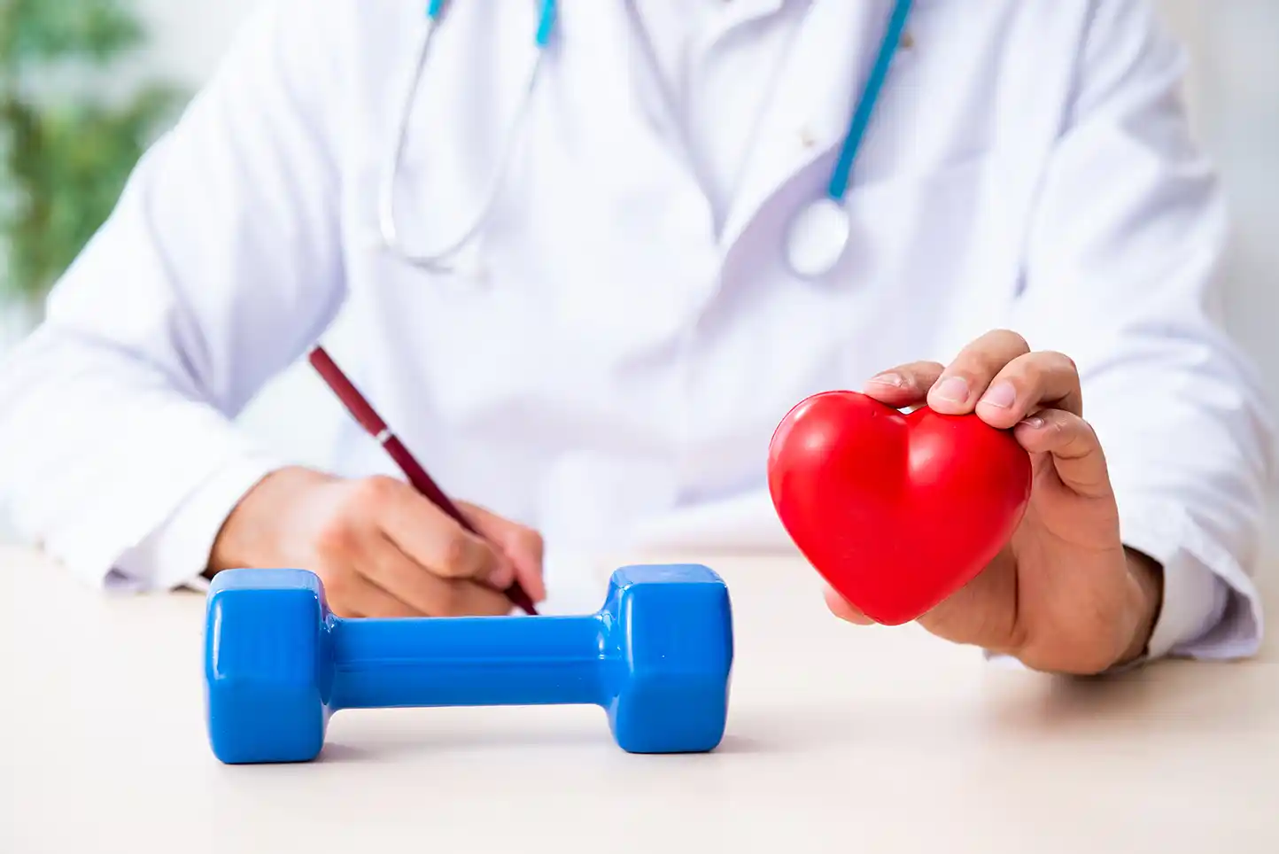 Médico de fondo, sosteniendo con una mano una pelota en forma de corazón rojo y con la otra mano sosteniendo una pluma, frente de él una pesa azul.