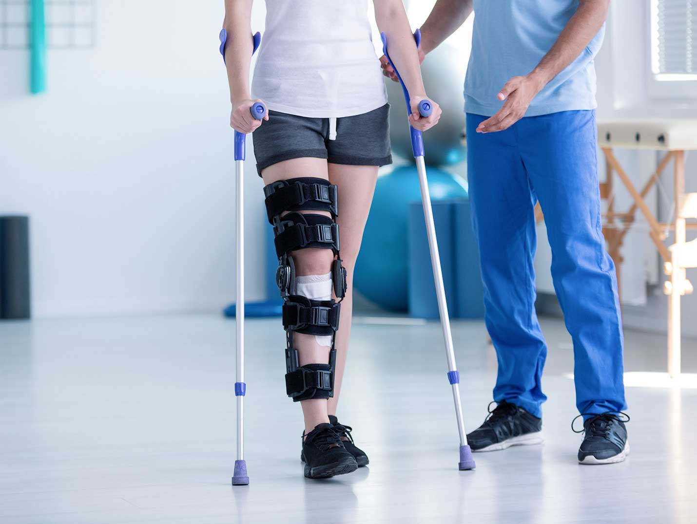 Paciente usando un soporte de rodilla negro en su pierna derecha y caminando con la ayuda de dos muletas. A su lado hay un médico