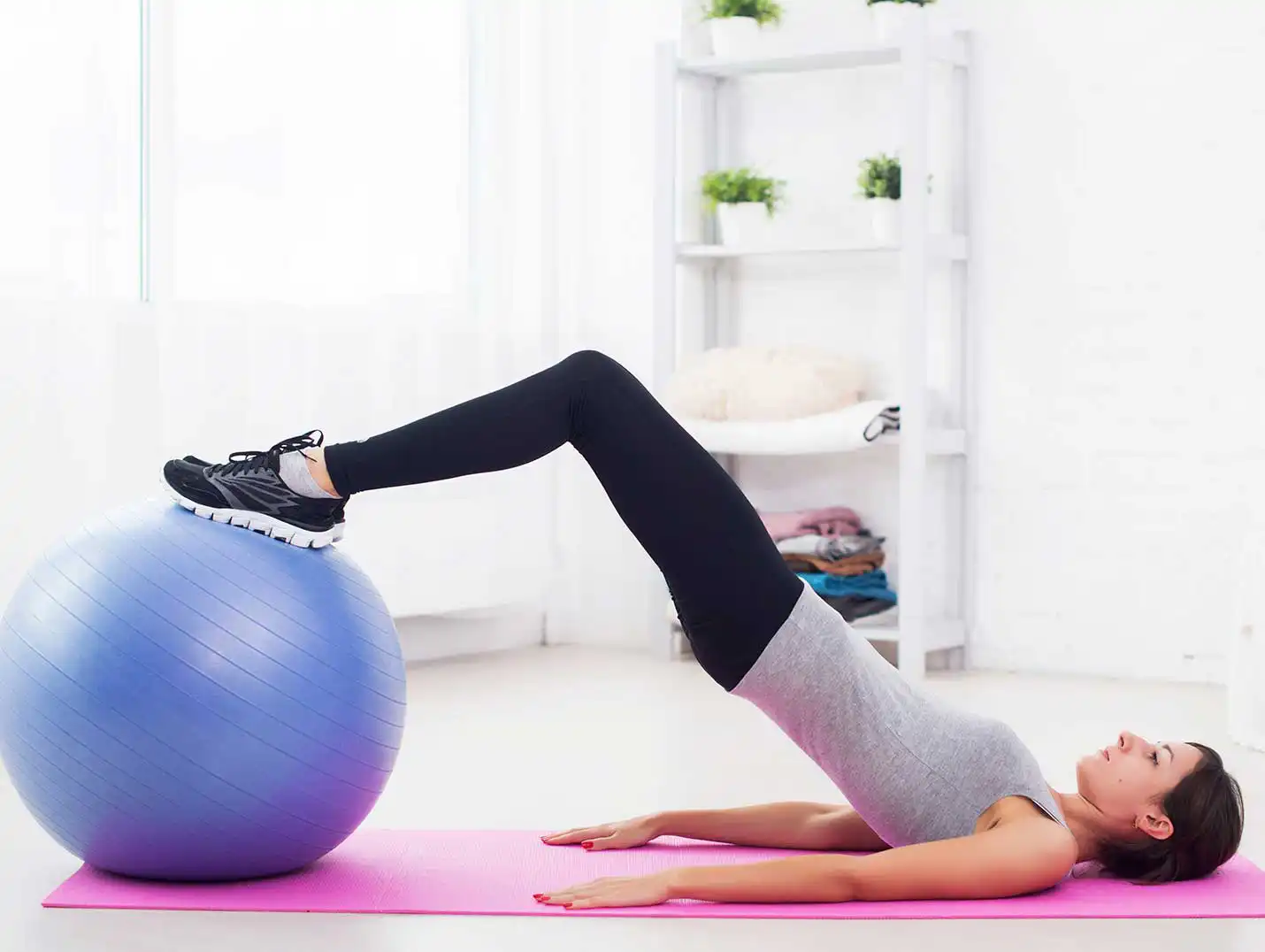 Mujer joven acostada sobre un tapete de yoga rosa, levantando la pelvis con la ayuda de una pelota para pilates azul