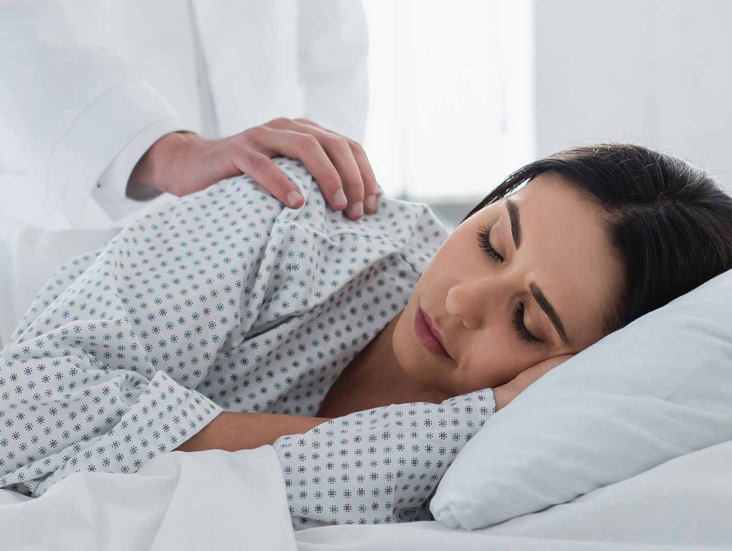 Paciente acostada y dormida en una cama con almohada y sábanas blancas. Un doctor está tocando su hombro con su mano