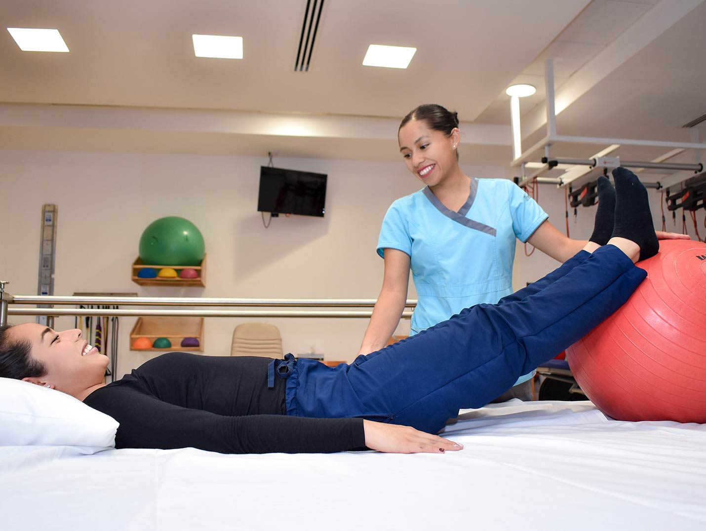 Paciente con playera negra y pantalon azul recostada sobre una cama y con los pies encima de una pelota junto a su rehabilitadora.