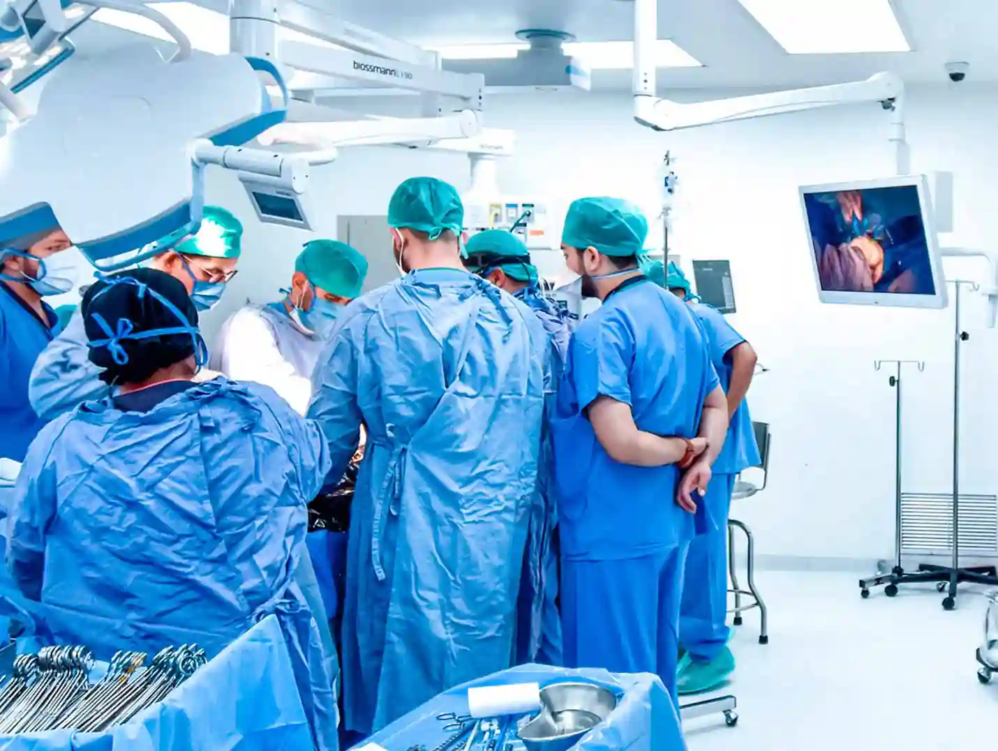 Ocho médicos con pijamas azules para cirugía, gorros verdes y cubrebocas, realizando un procedimiento quirúrgico