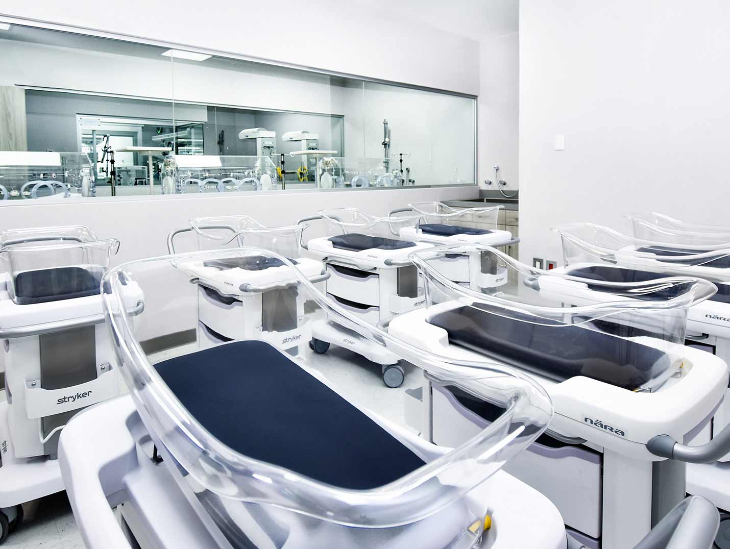 Area de neonatología con paredes blancas y 8 cuneros. Al fondo un cristal donde se ve la sala de incubadoras.