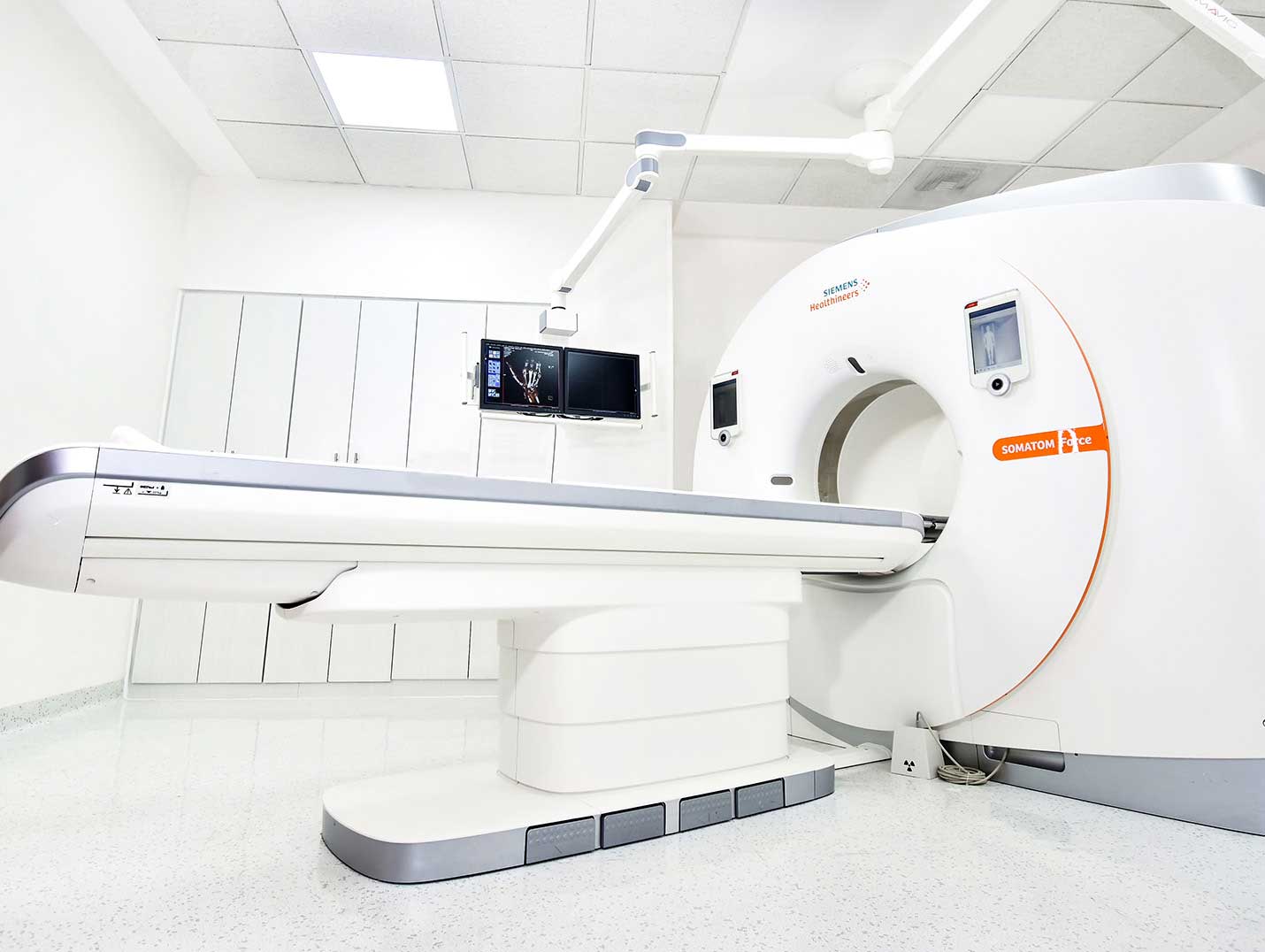 Cuarto amplio y blanco con equipo de tomografía y pantallas táctiles a un costado