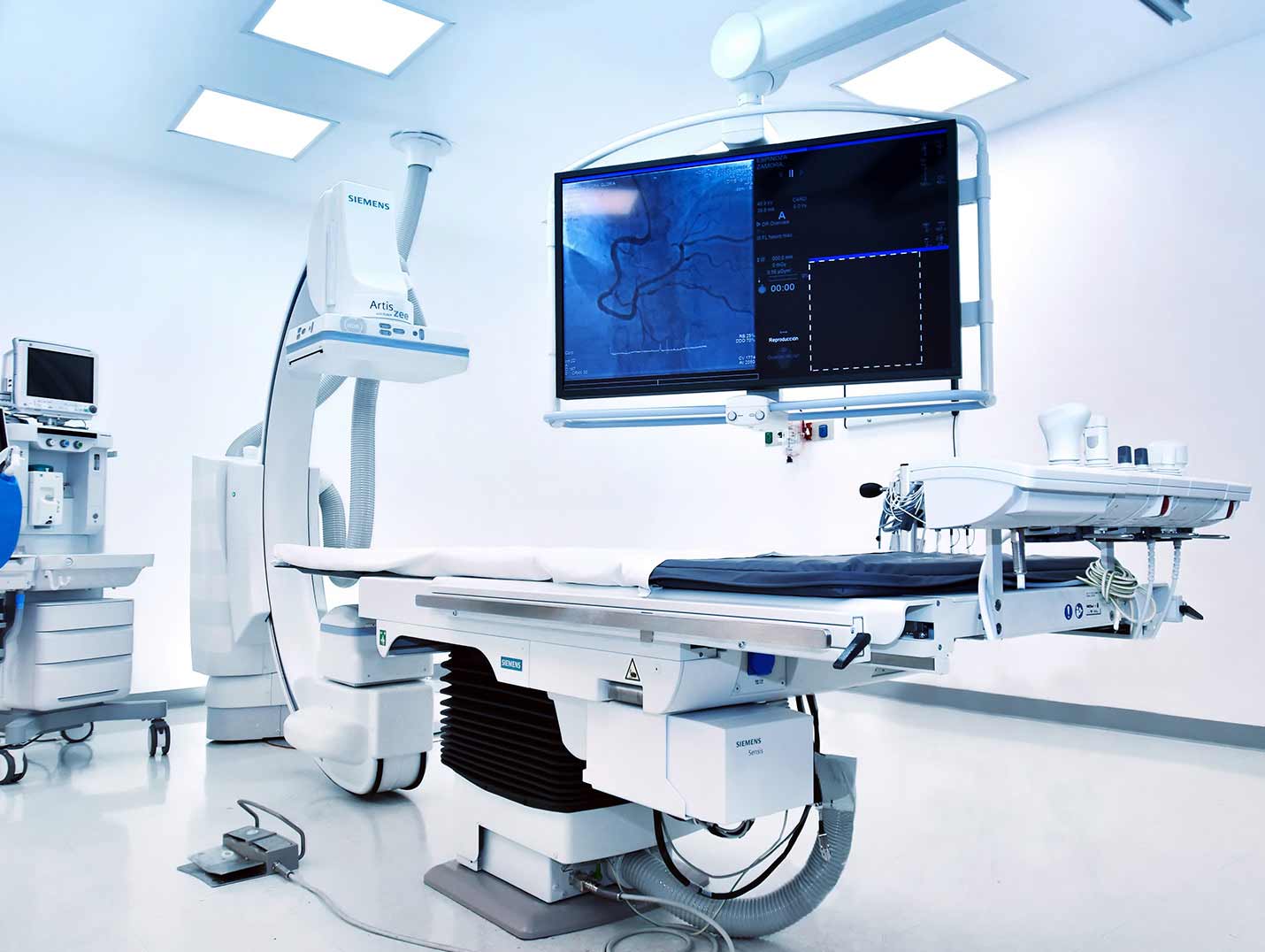 Equipo de hemodinamia de color blanco con pantalla grande mostrando un estudio vascular en un cuarto con luces azules.