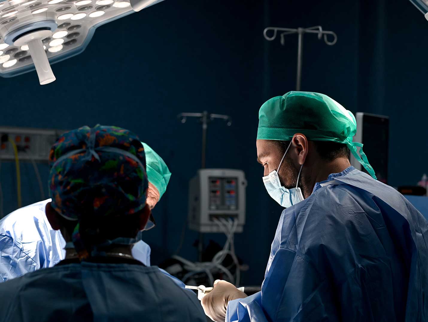 Tres médicos con pijamas azules para cirugía, gorros, cubrebocas y guantes blancos, realizando un procedimiento quirúrgico