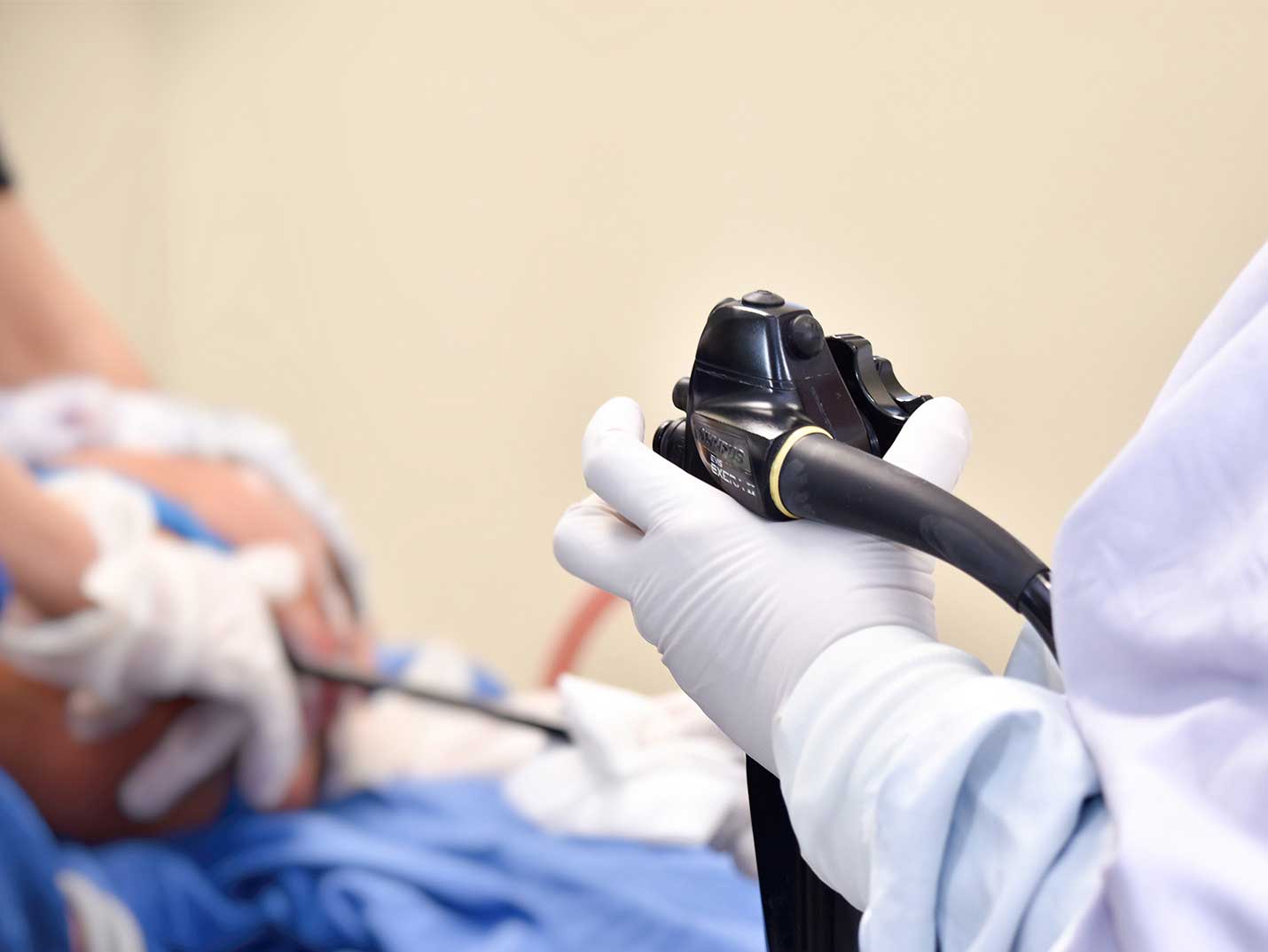 Mano de un médico con un guante blanco, sosteniendo un endoscopio negro. Al fondo está otro médico sujetando la cabeza de un paciente