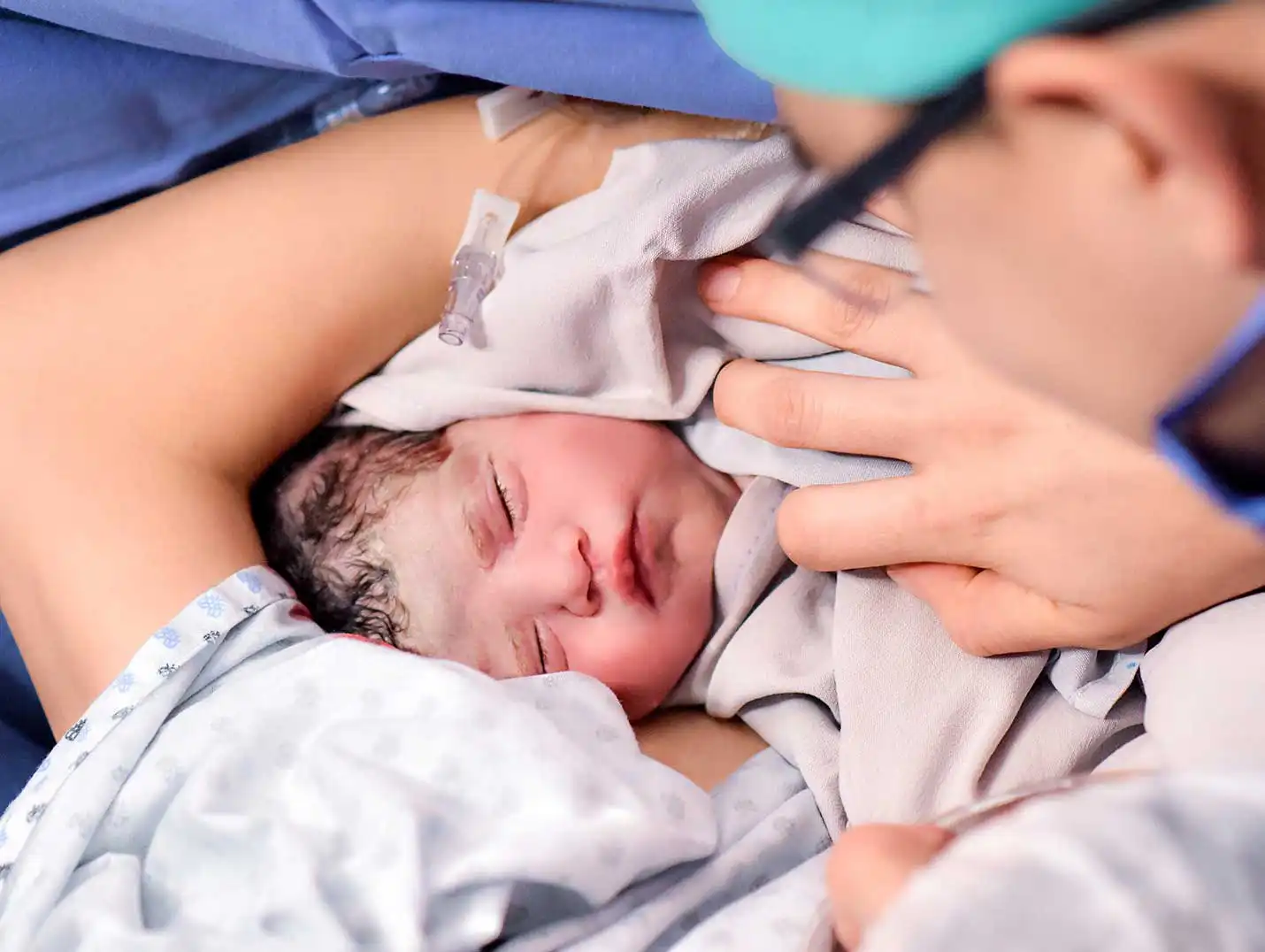 Mamá vestida con una bata para paciente, cargando a su bebé recién nacido que está dormido y envuelto en una manta