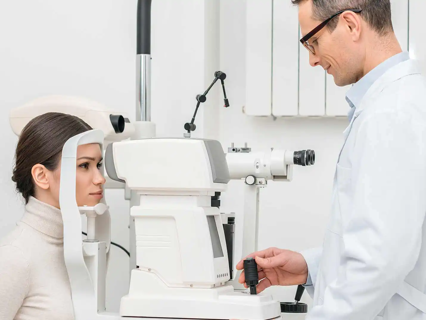 Doctor con bata blanca realizando un examen oftalmológico a una paciente que viste un suéter de cuello alto blanco