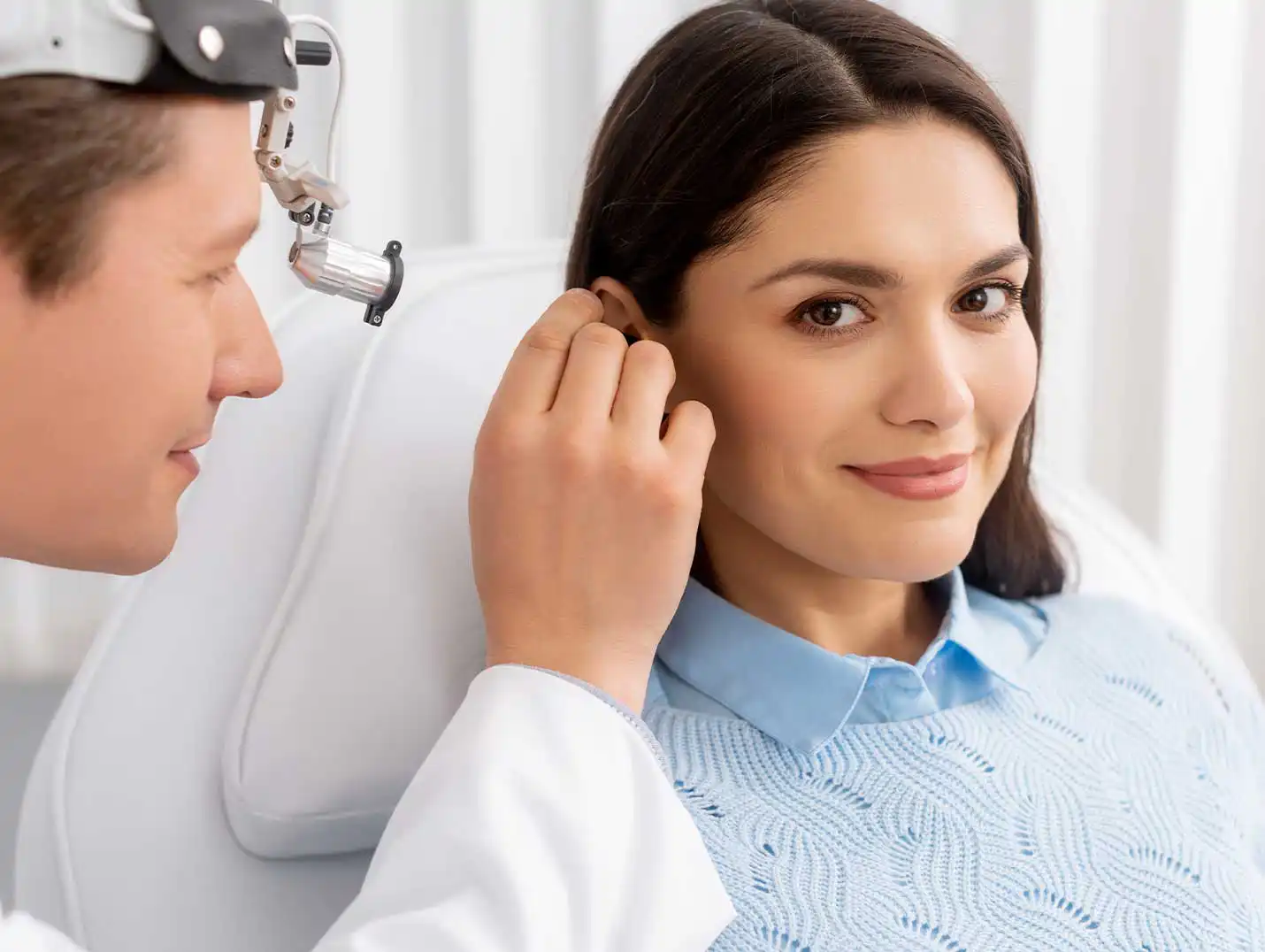 Doctor con bata blanca realizando un examen auditivo a una paciente que está sentada y viste un suéter azul