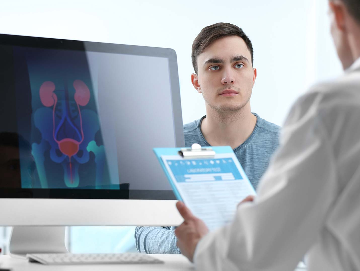 Doctor sosteniendo una tabla con clip azul, mientras un paciente lo mira detenidamente. Hay un monitor mostrando el sistema urinario