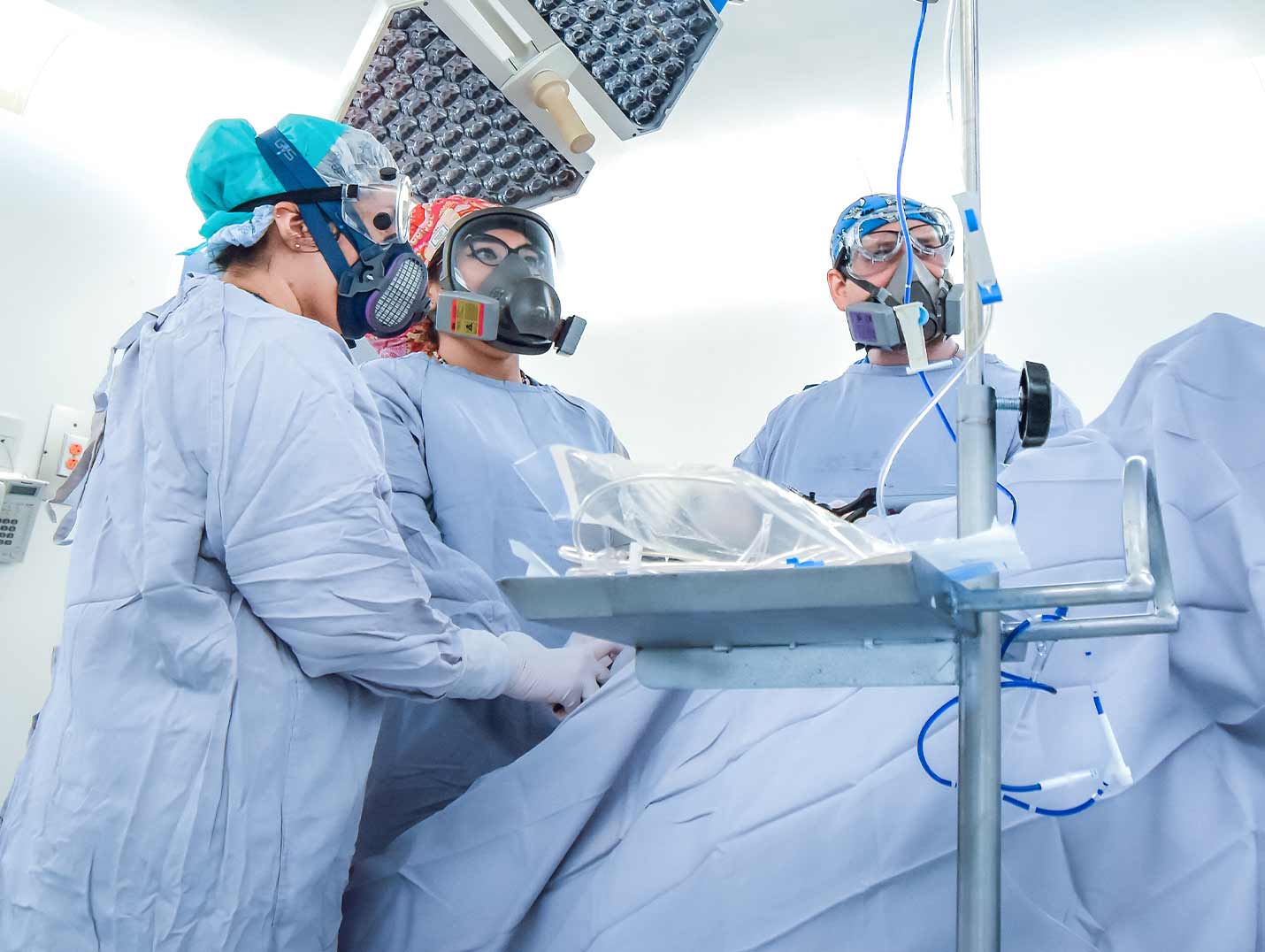 Tres médicos en un quirófano vestidos con pijamas azules para cirugía y usando respiradores, realizando una cirugía