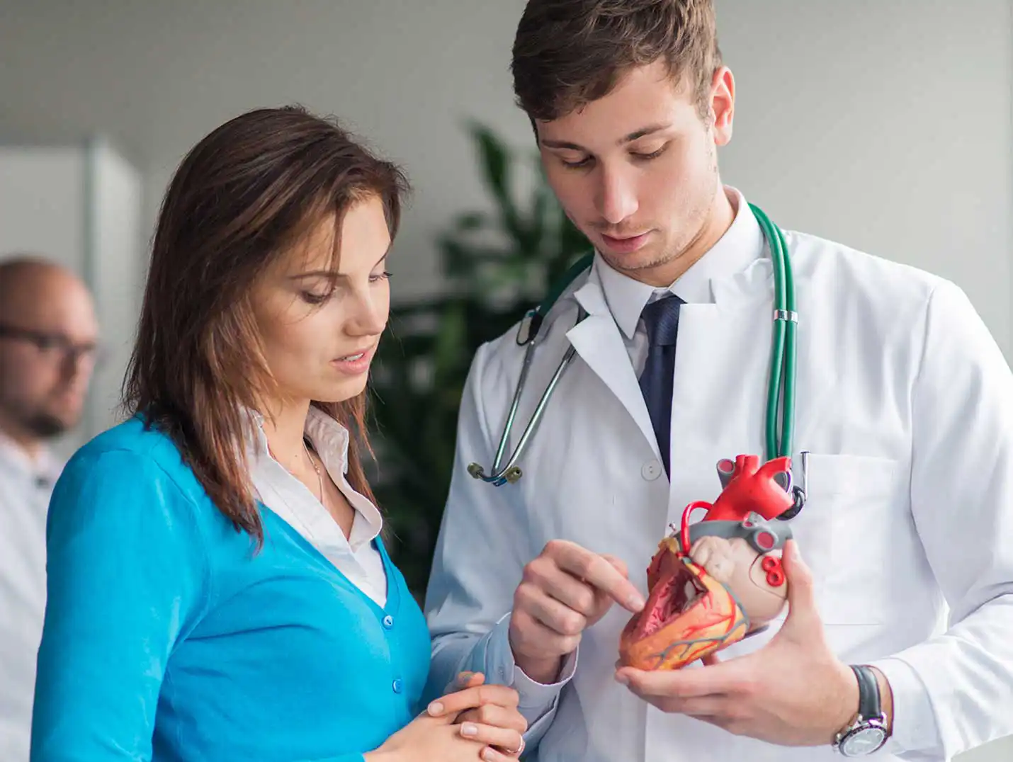 Doctor con bata blanca y un estetoscopio verde alrededor de cuello, mostrándole a una paciente un prototipo del corazón