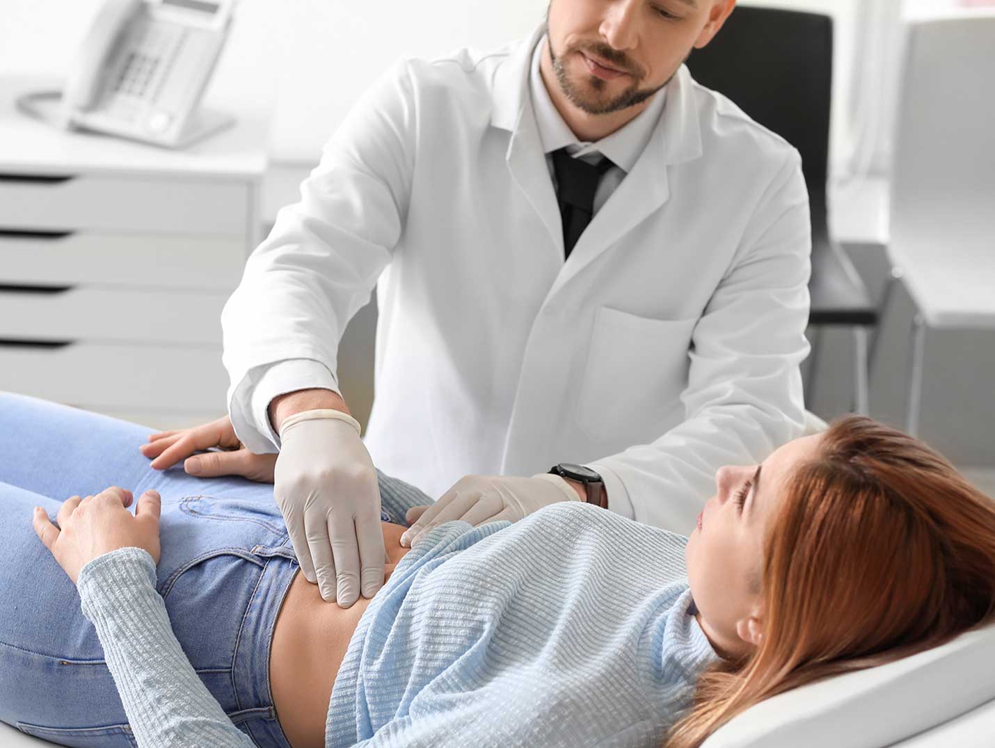 Doctor con bata y guantes blancos palpando el abdomen de una paciente que está acostada en una mesa de exploración