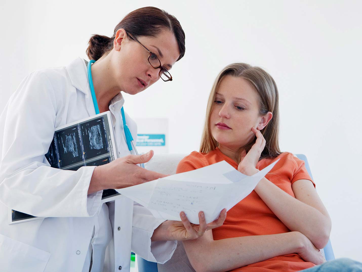 Doctora con bata blanca mostrándole los resultados de un estudio a una paciente que está sentada y viste una blusa naranja