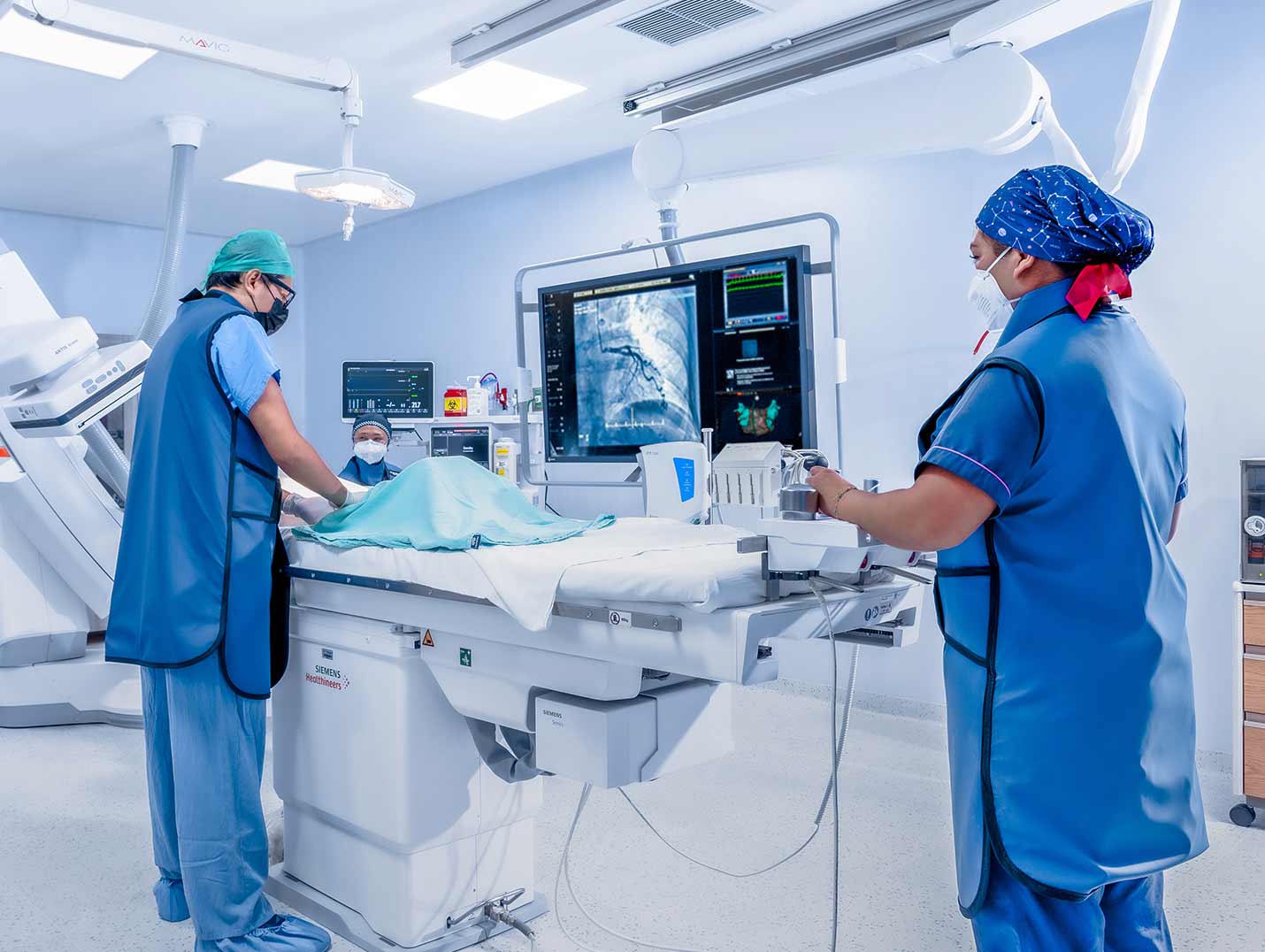 Tres médicos en la sala de hemodinamia vestidos con pijamas azules para cirugía y gorros. El monitor muestra una radiografía de tórax