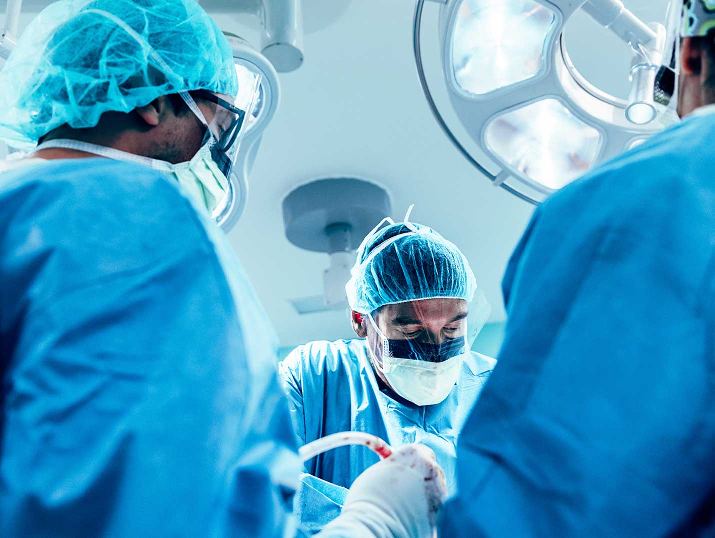 Tres médicos con pijamas azules para cirugía, cofias, cubrebocas y guantes blancos, realizando una cirugía