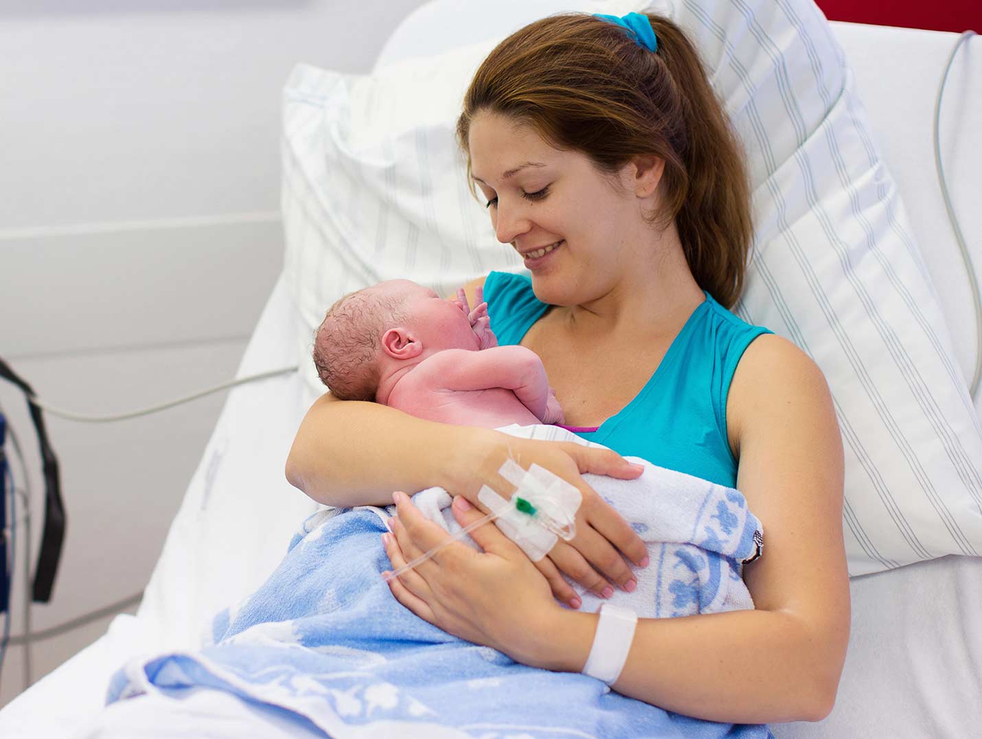 Una mamá recostada en una cama hospitalaria con un suero intravenoso puesto, cargando y mirando a su bebé recién nacido