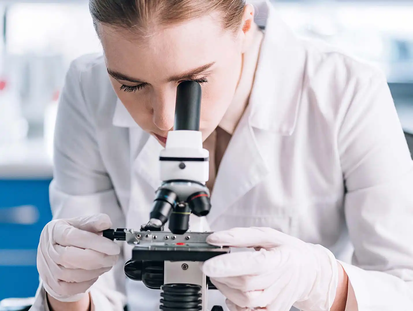 Doctora con bata y guantes blancos, acomodando y analizando una muestra de laboratorio con un microscopio