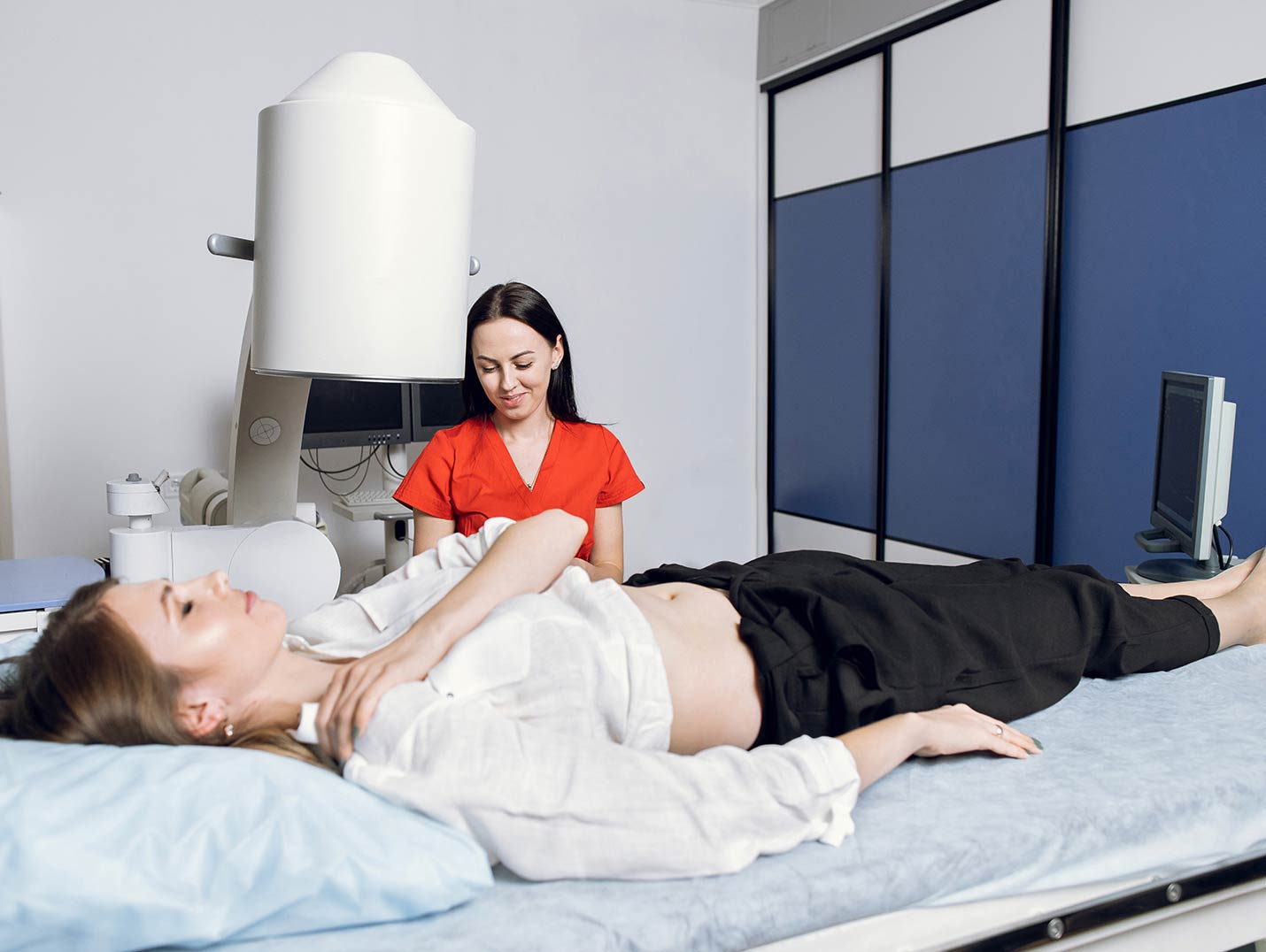 Paciente acostada con los ojos cerrados en la camilla del litotriptor. Una doctora con uniforme naranja está sentada a su lado