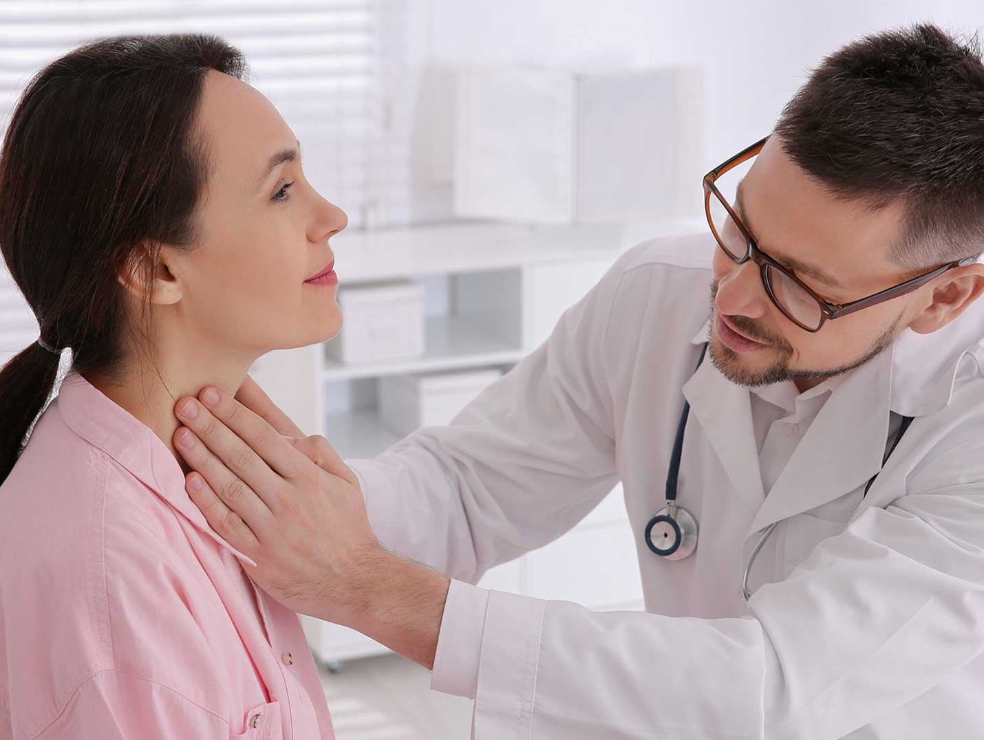 Doctor con bata blanca, lentes y un estetoscopio alrededor del cuello, palpando la garganta de una paciente con blusa rosa