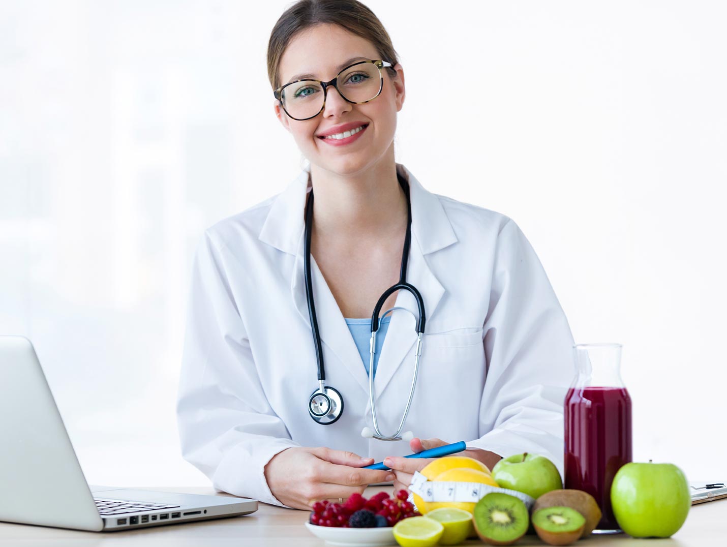 Doctora sonriendo, vestida con una bata blanca. A su lado hay una MacBook, diversas frutas, un jugo y una cinta métrica