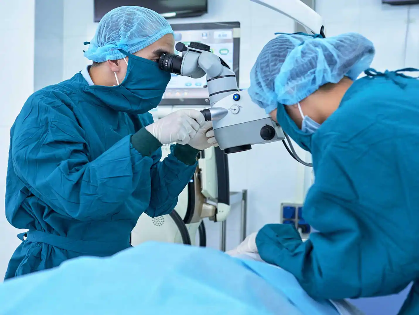 Dos médicos vestidos con pijamas azules para cirugía, cofias y guantes blancos, realizando una cirugía láser