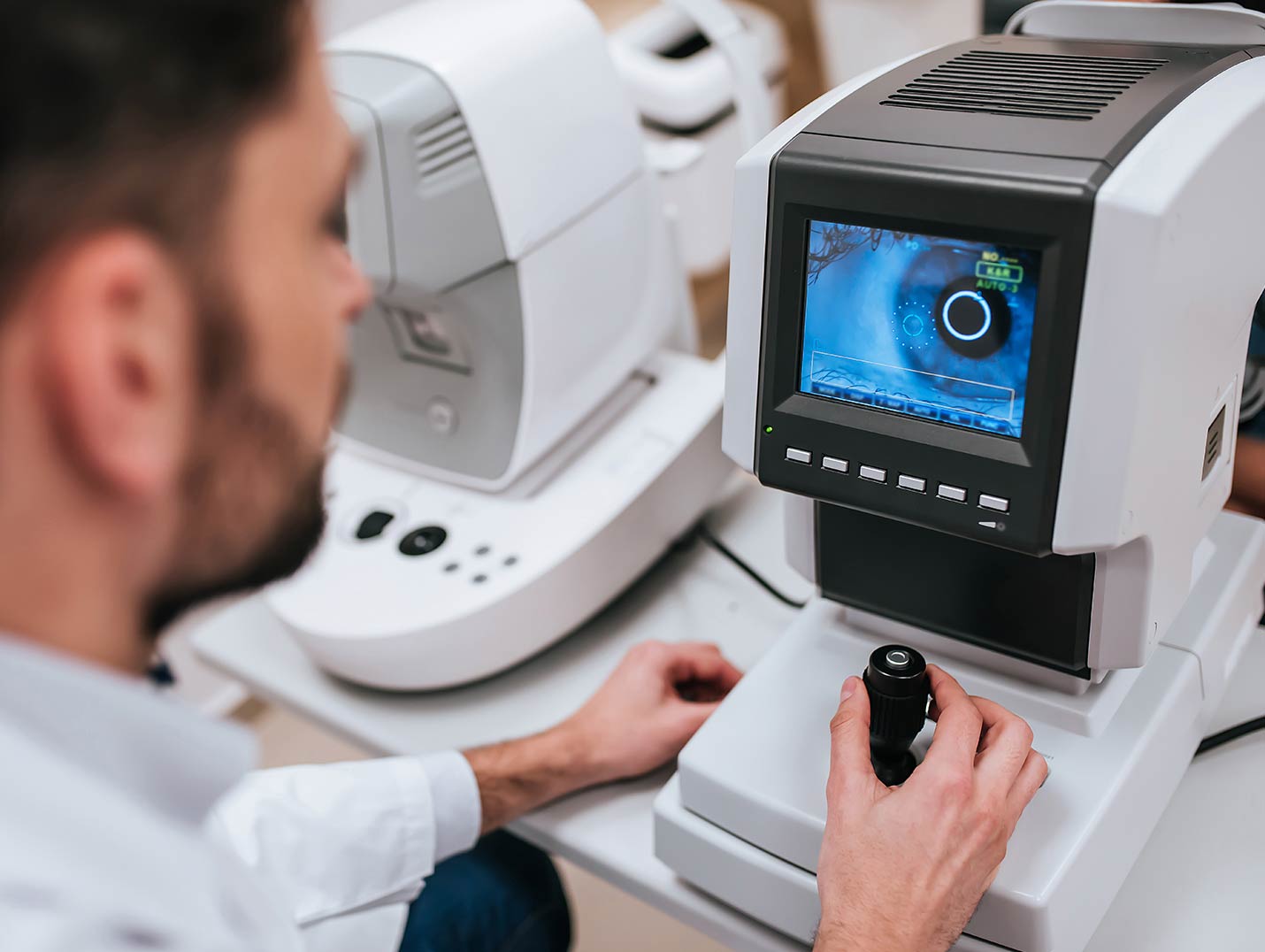 Médico con bata blanca explorando la retina de un paciente con un autorefractómetro blanco