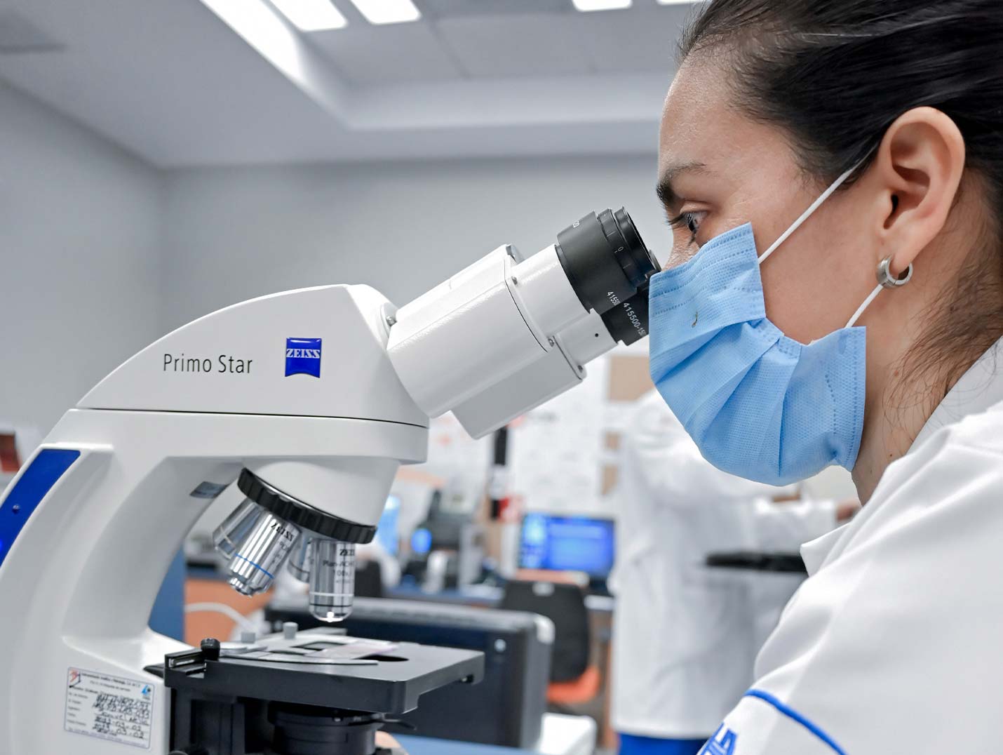 Doctora de Hospital Angeles con bata blanca y cubrebocas, analizando una muestra de laboratorio con el microscopio