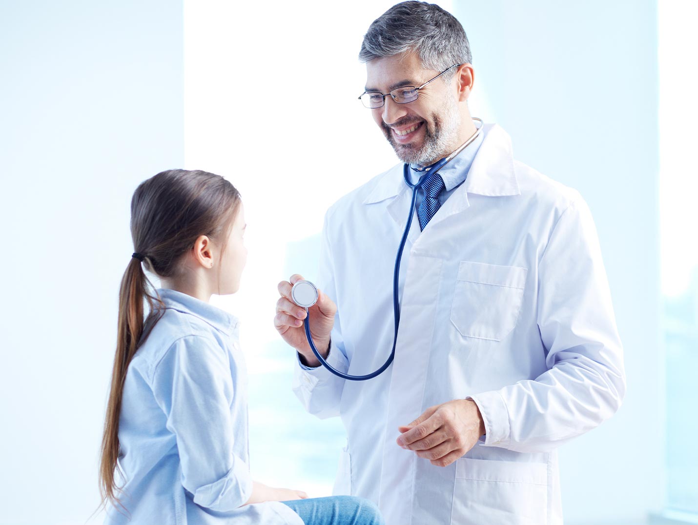 Doctor con bata y guantes blancos sonriendo y sosteniendo un estetoscopio. En frente de él hay una niña pequeña sentada